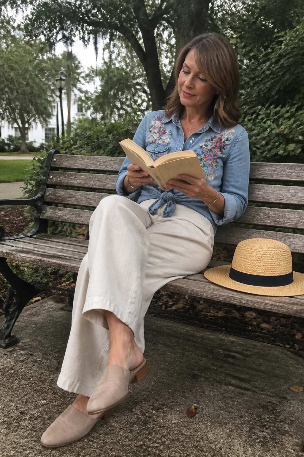 Woman over 50 wearing embroidered light blue chambray shirt tied at the waist with cream wide leg pants and beige suede mules while seated on a bench holding an open book with a straw hat beside her