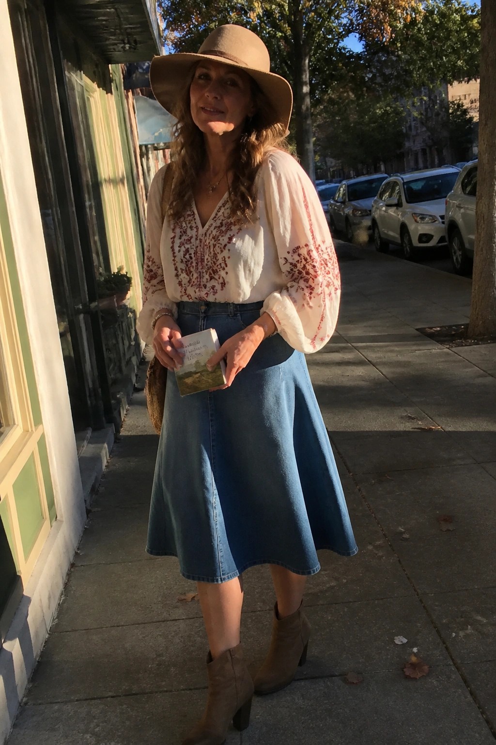 Woman in wide-brim hat, embroidered blouse, and denim skirt holding book on sidewalk