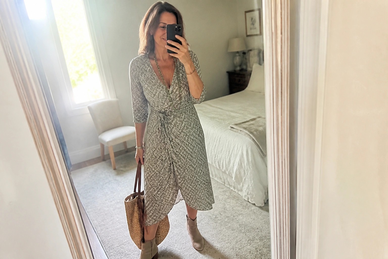 Woman in brown floral wrap dress holding straw bag in bedroom mirror selfie
