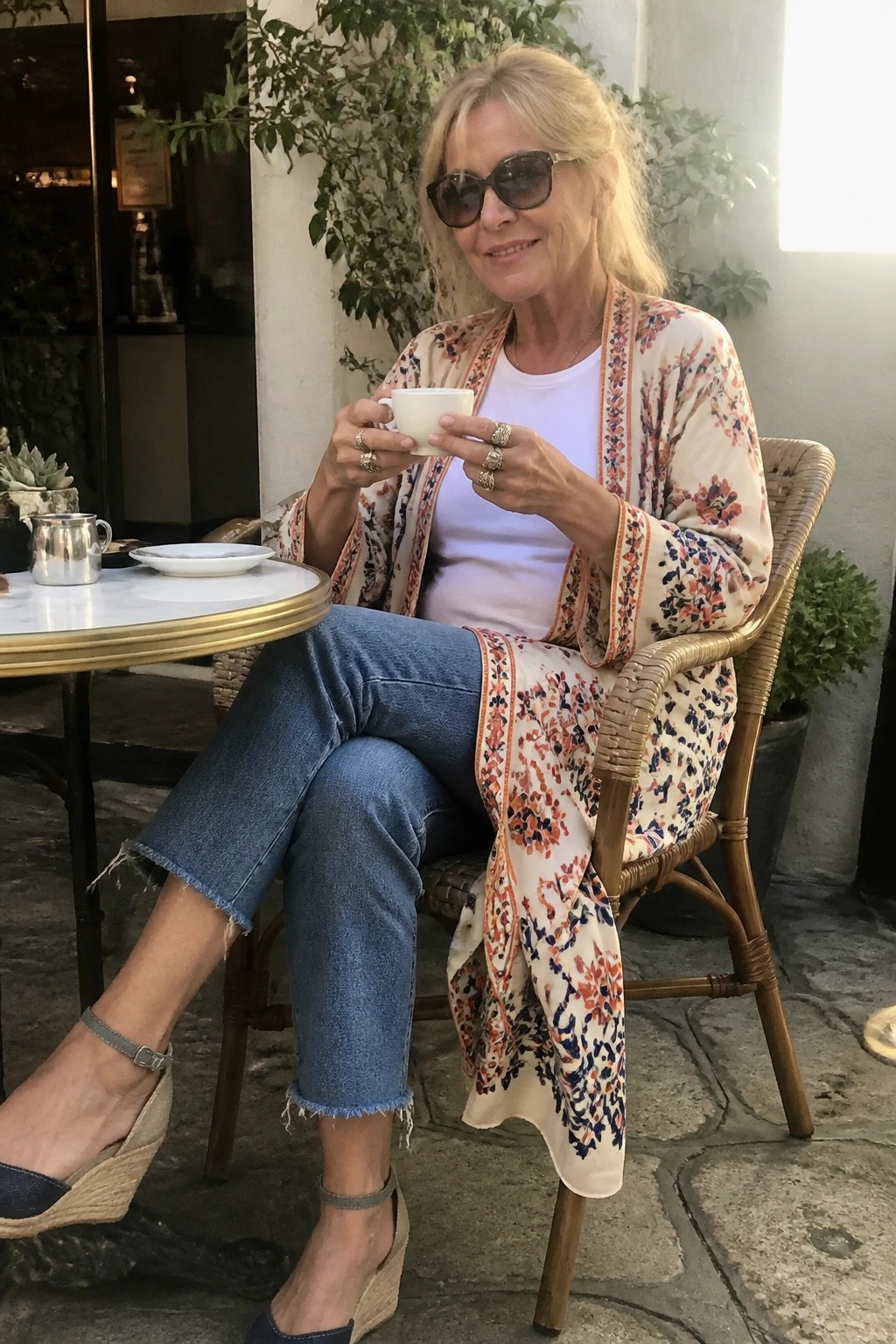 Woman over 50 in floral print kimono cardigan over white tee and cropped blue jeans with frayed hems paired with wedge espadrilles and rings while seated outdoors holding a coffee cup.