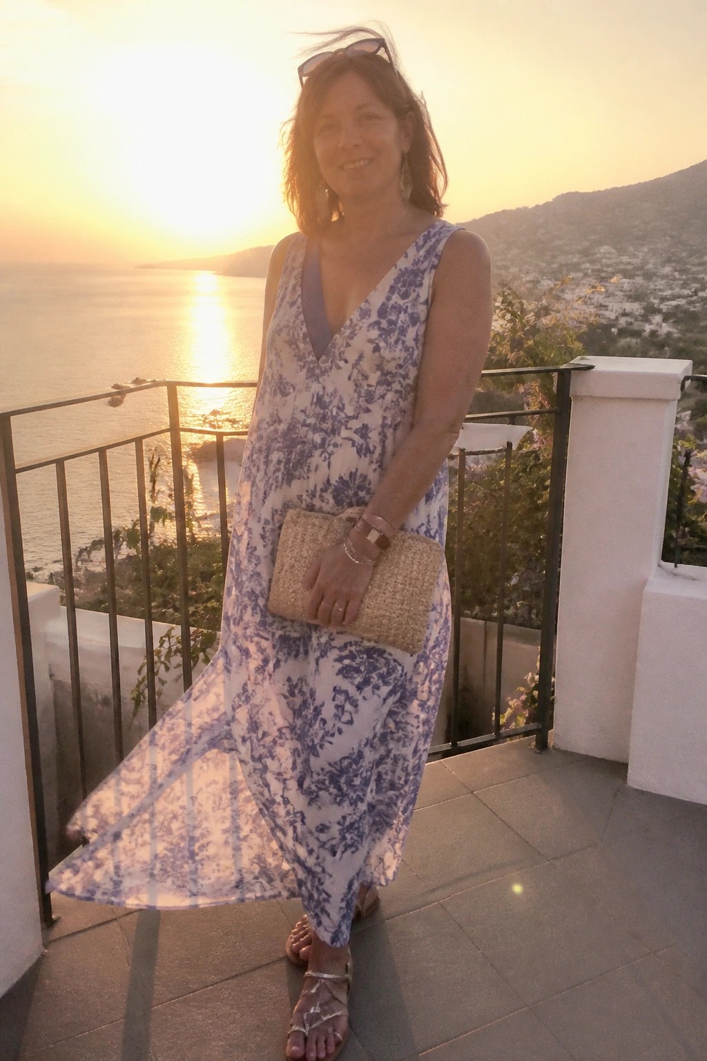 Woman in floral dress holding clutch on sunset balcony overlooking sea