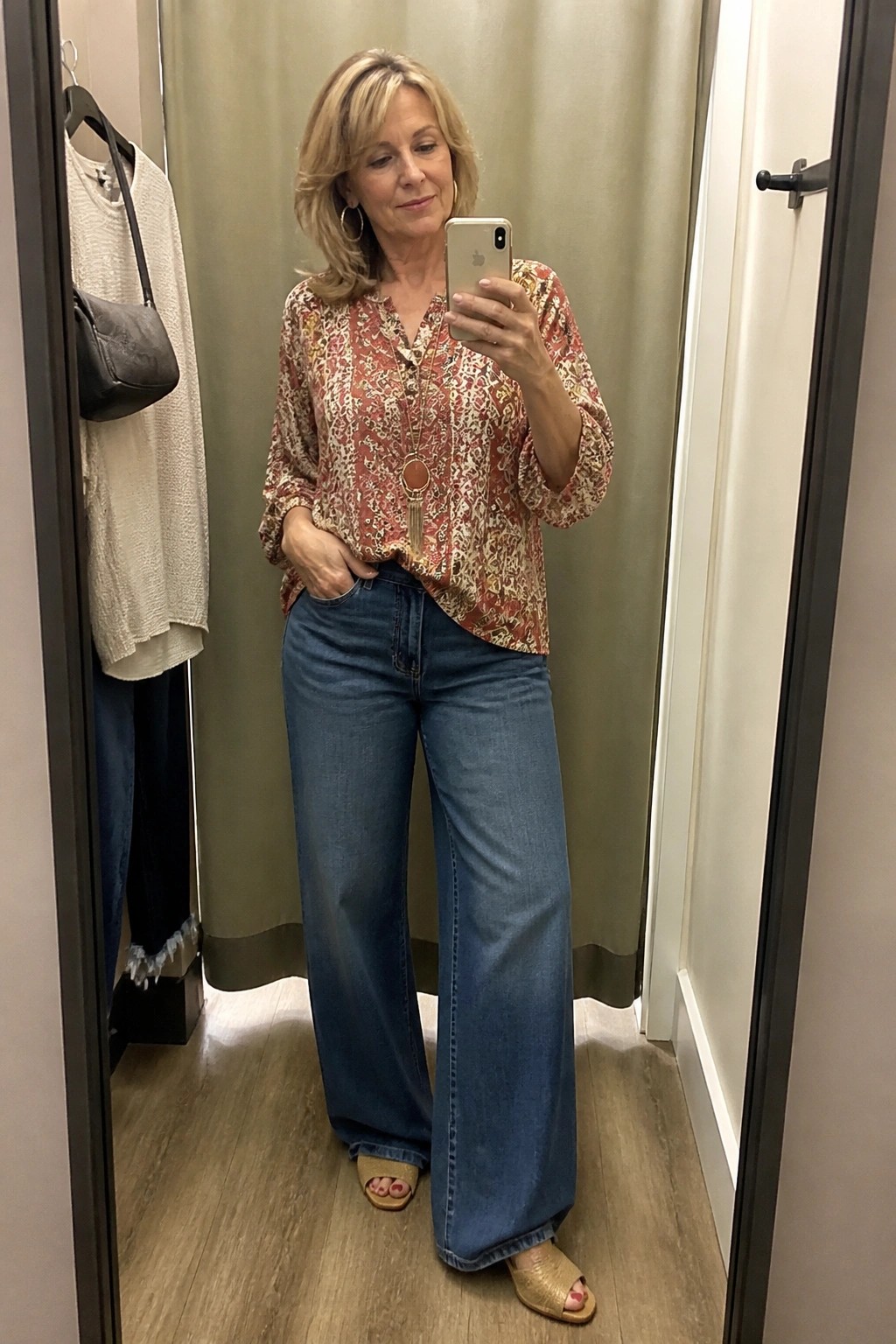 Woman in patterned blouse and wide-leg jeans taking mirror selfie in fitting room