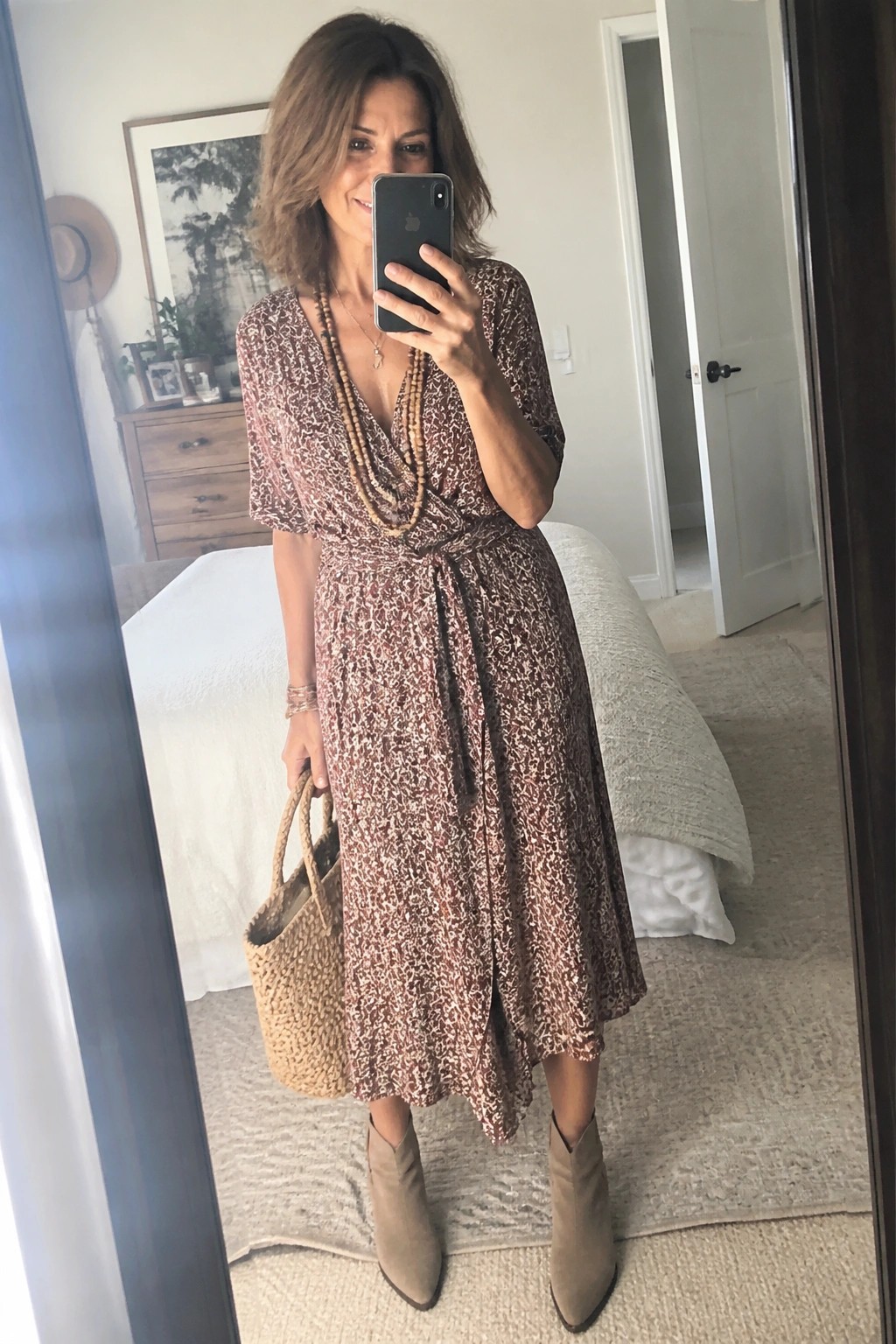 Woman in brown floral wrap dress holding straw bag in bedroom mirror selfie