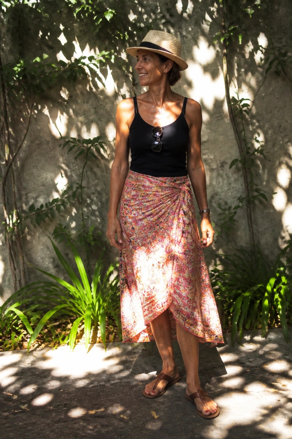 Woman over 50 in black tank top tucked into a colorful patterned wrap skirt with straw hat, brown flip flops, and sunglasses tucked into the neckline standing against a stone wall.