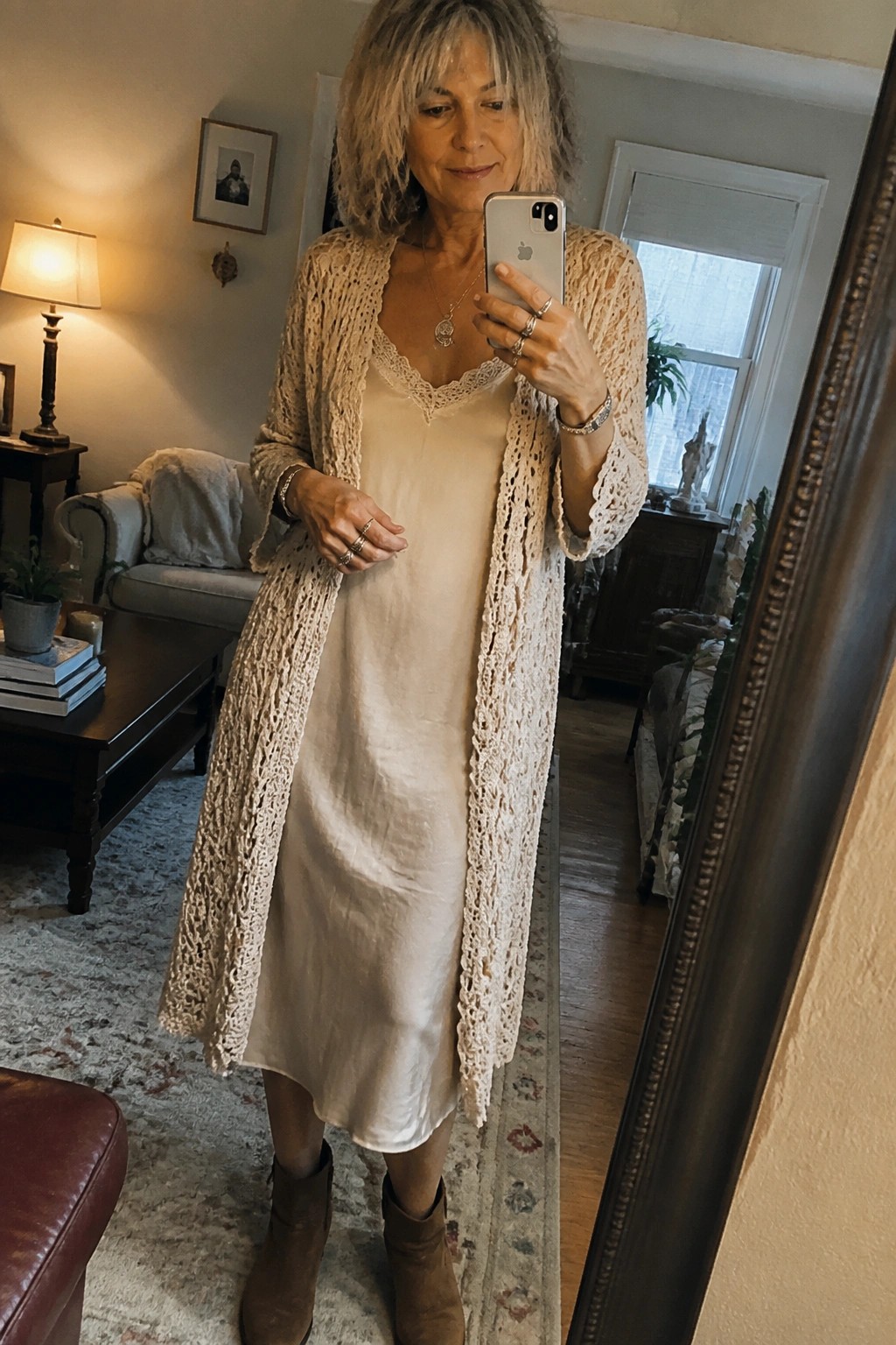 Woman with gray hair in cream slip dress and beige crochet cardigan taking mirror selfie