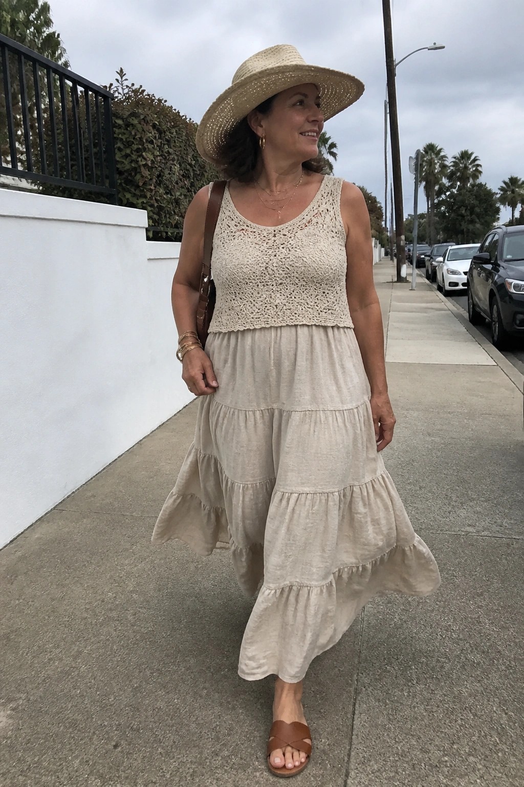 Smiling woman in straw hat and tiered beige dress walking on sidewalk.