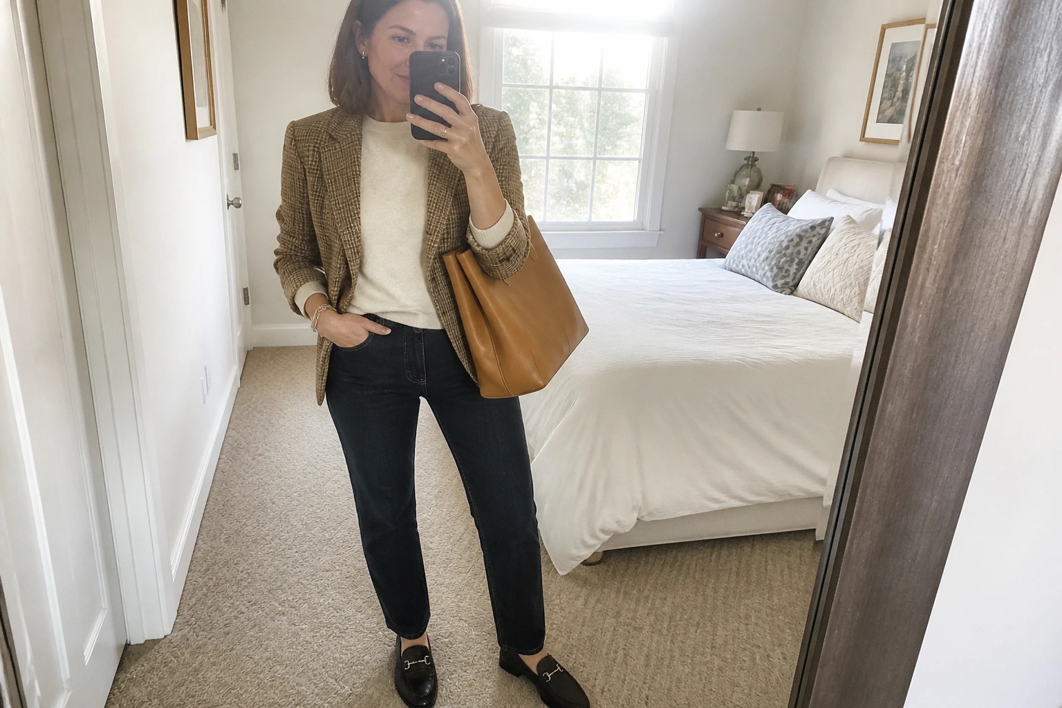 Woman wearing a brown tweed blazer over a cream sweater with dark straight leg jeans black loafers and a brown leather tote bag while holding a phone for a mirror selfie.