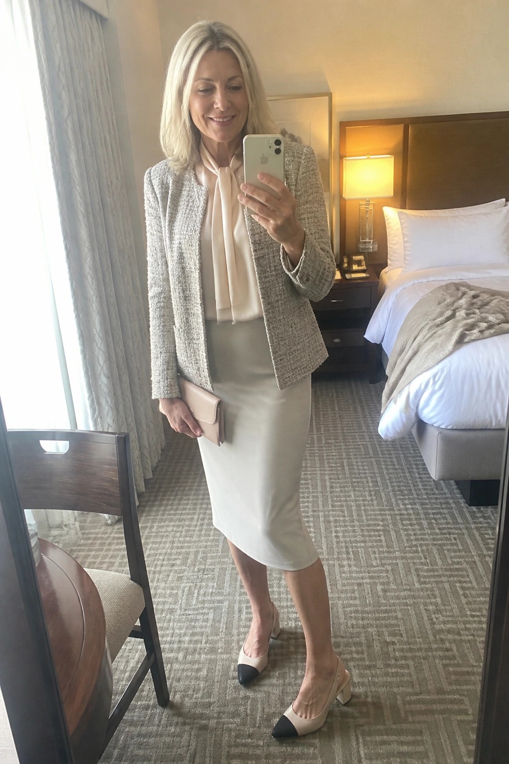 Blonde woman in tweed blazer and pencil skirt taking mirror selfie in hotel room