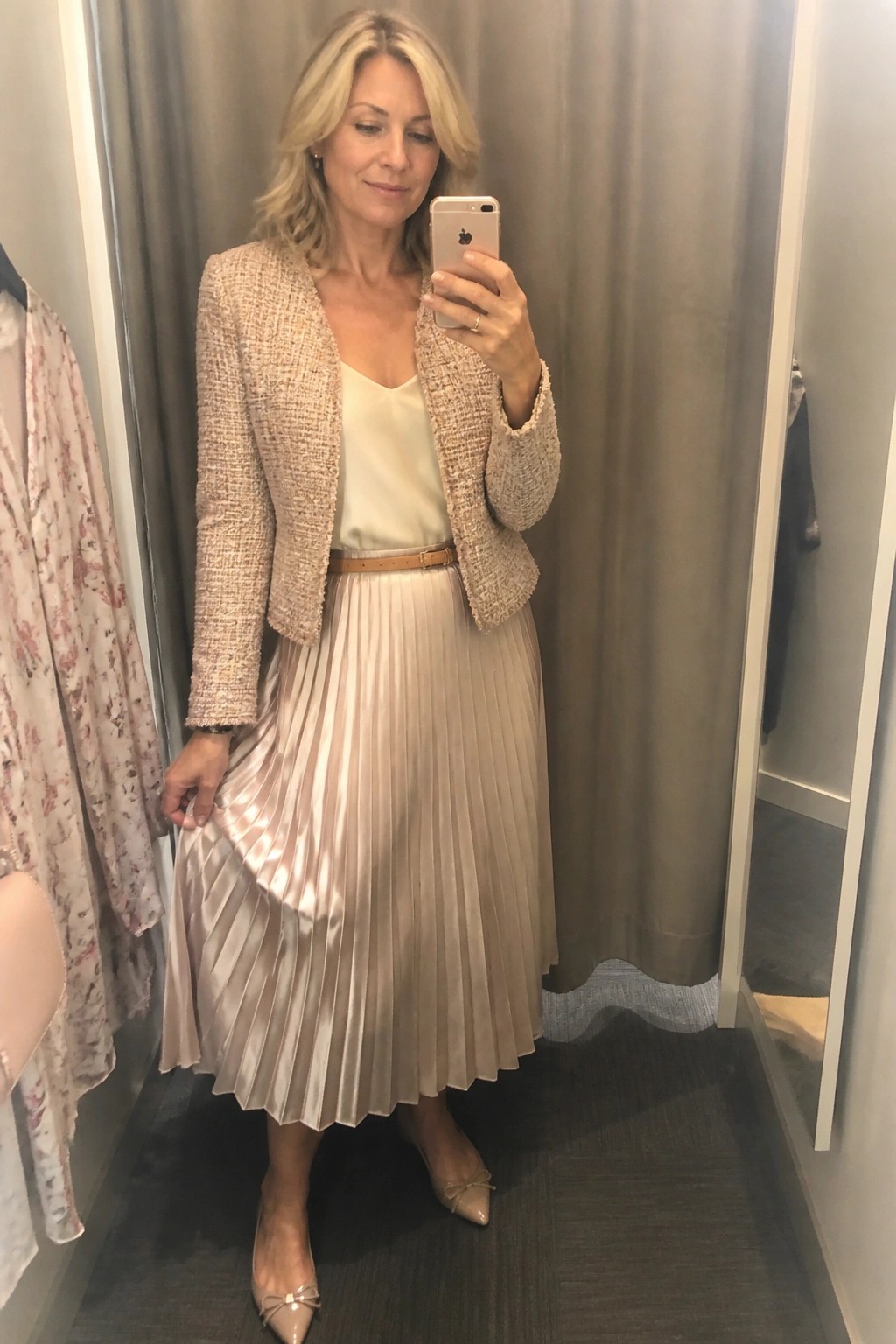 Blonde woman in beige tweed jacket and pleated satin skirt taking mirror selfie in fitting room