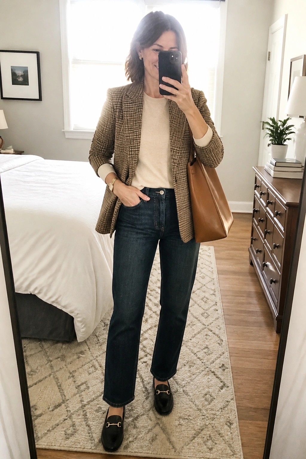 Woman wearing a brown tweed blazer over a cream sweater with dark straight leg jeans black loafers and a brown leather tote bag while holding a phone for a mirror selfie.