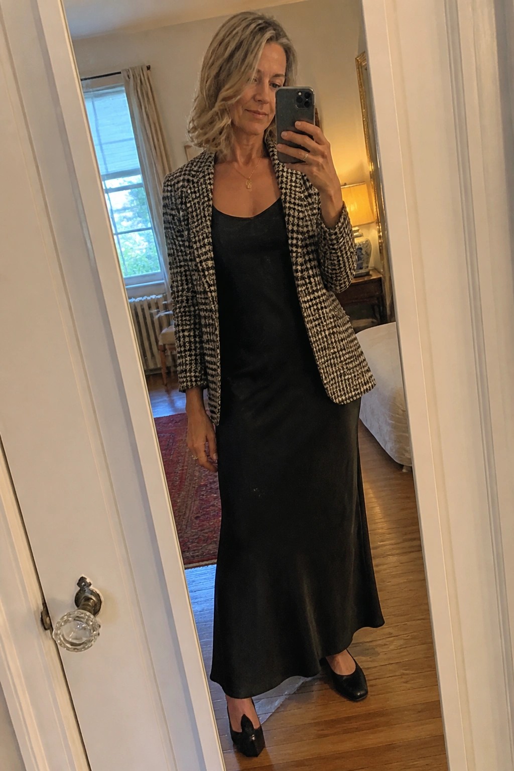 Woman in black slip dress and houndstooth blazer taking mirror selfie in bedroom