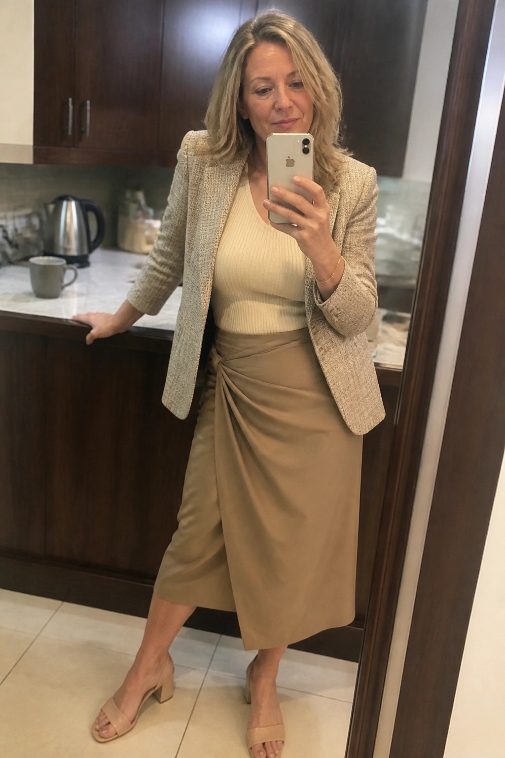 Blonde woman in beige blazer and taupe wrap skirt taking mirror selfie in kitchen
