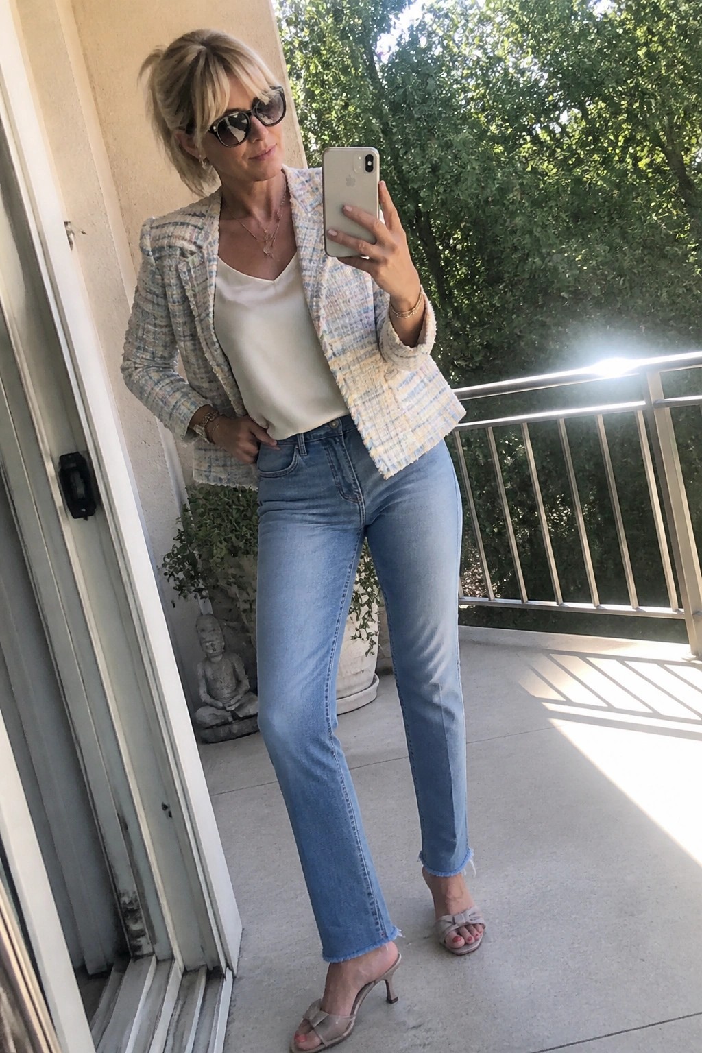 Woman over 50 wearing a multicolor tweed blazer over a white camisole with light blue straight leg jeans that have frayed hems, paired with nude heeled sandals and layered gold necklaces.