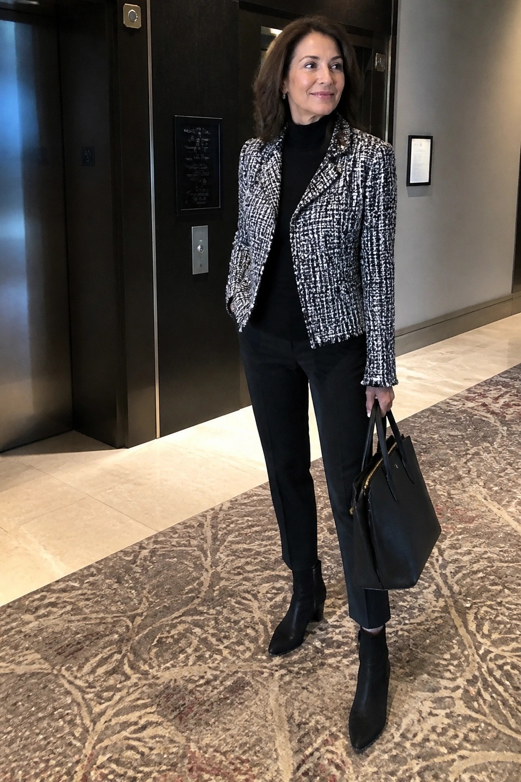 Woman in black-and-white tweed jacket, turtleneck, pants, and boots holding black handbag.
