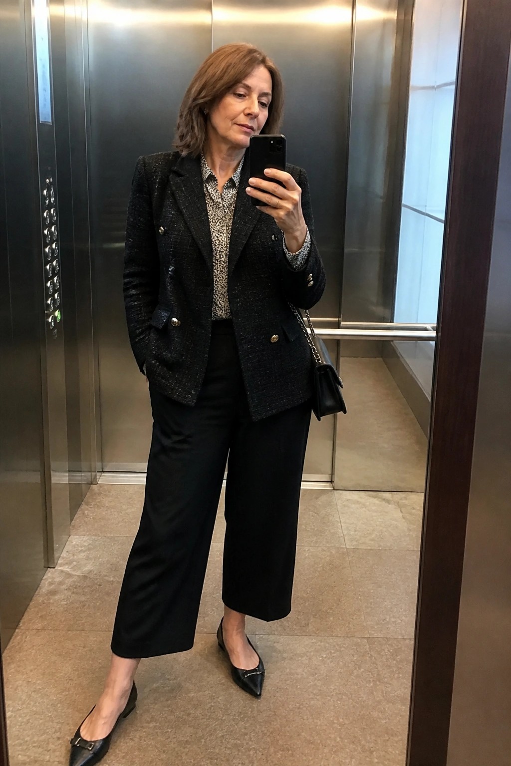 Woman in black tweed blazer and cropped pants taking selfie in elevator