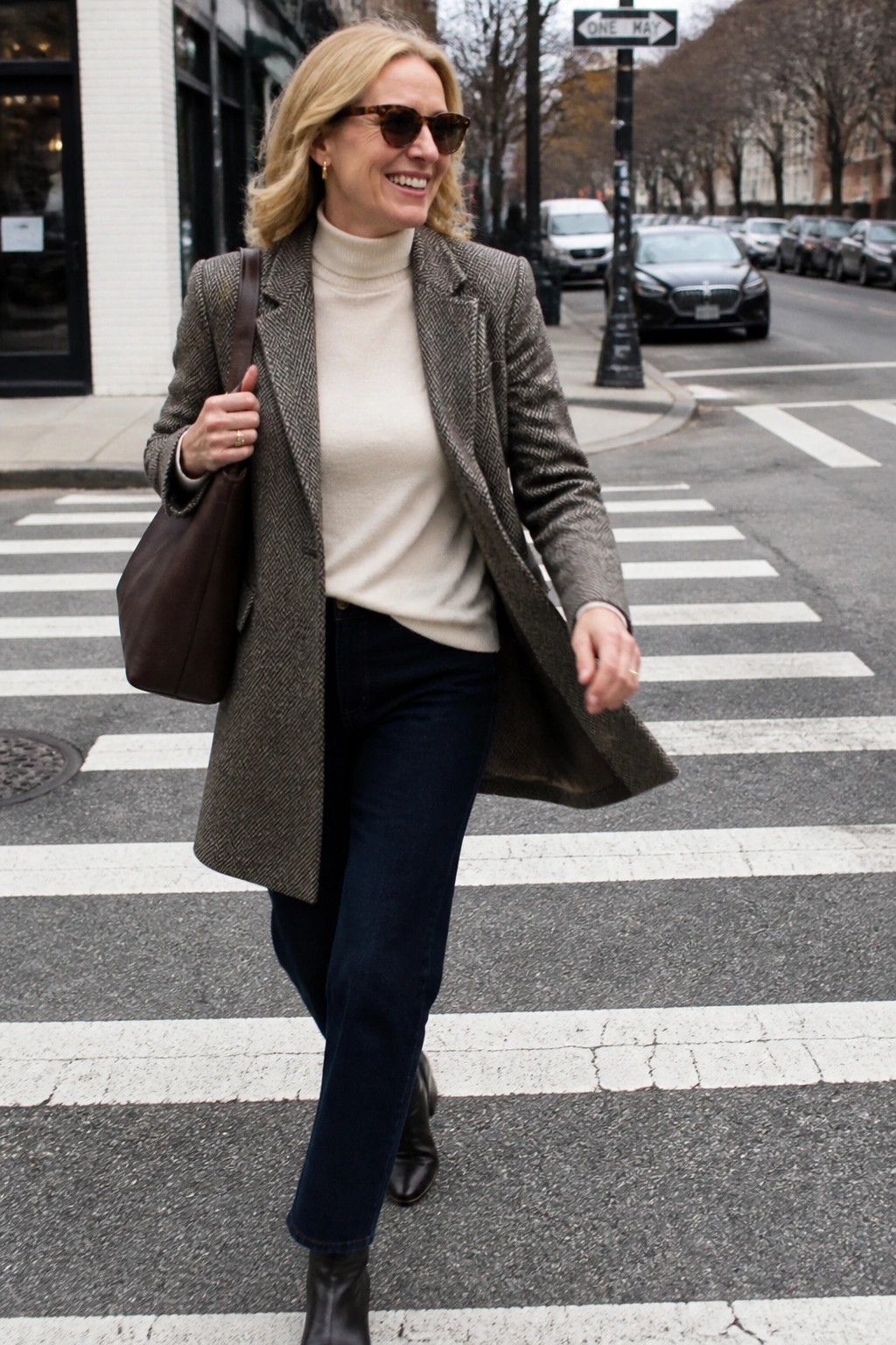 Smiling blonde woman in tweed blazer and turtleneck crossing urban crosswalk with tote bag