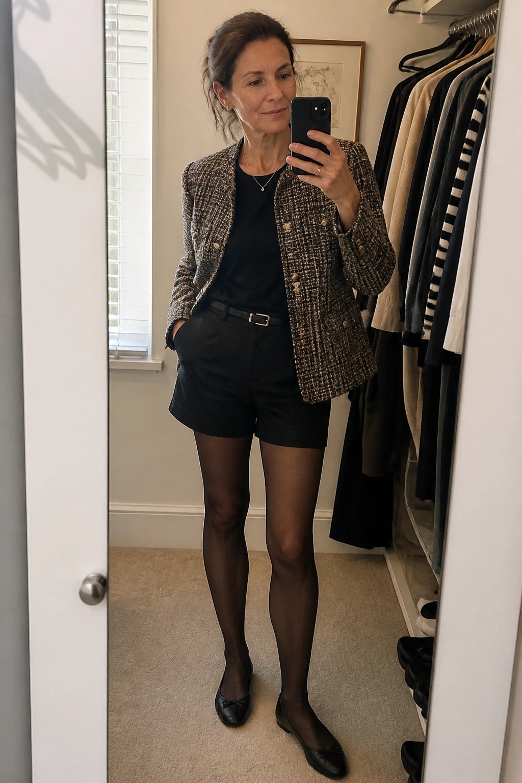 Woman in tweed jacket and black shorts taking mirror selfie in closet