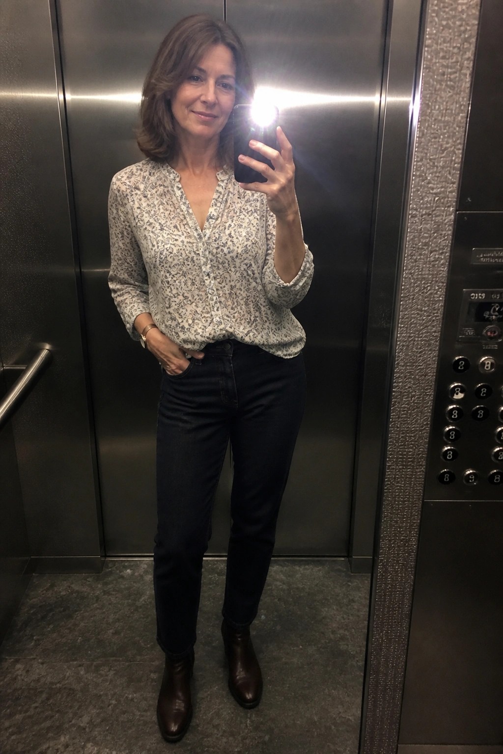 Woman in floral blouse and dark jeans taking flash selfie in elevator