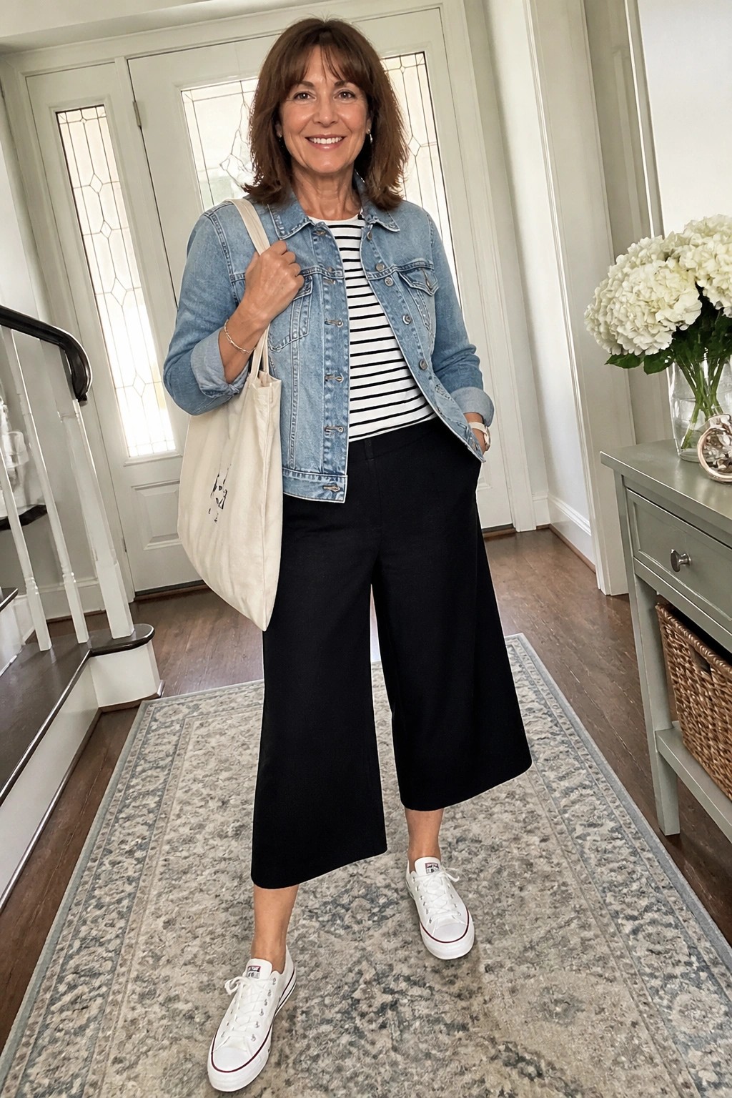 Woman in denim jacket over stripes with black culottes, white sneakers, holding tote bag.