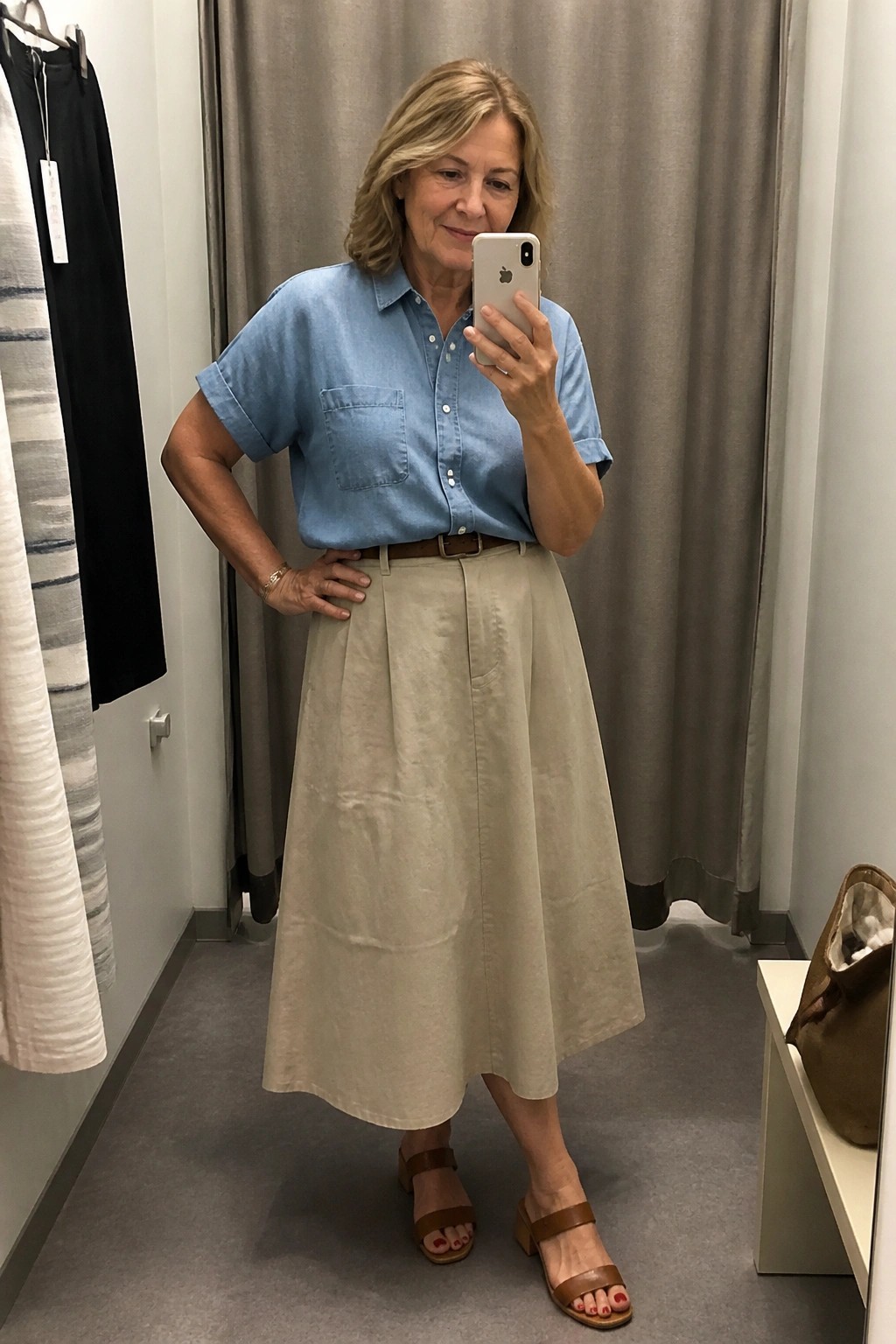 Woman over 50 wearing light blue short sleeve chambray shirt tucked into beige midi skirt with brown belt and matching brown sandals while taking a mirror selfie in a fitting room.