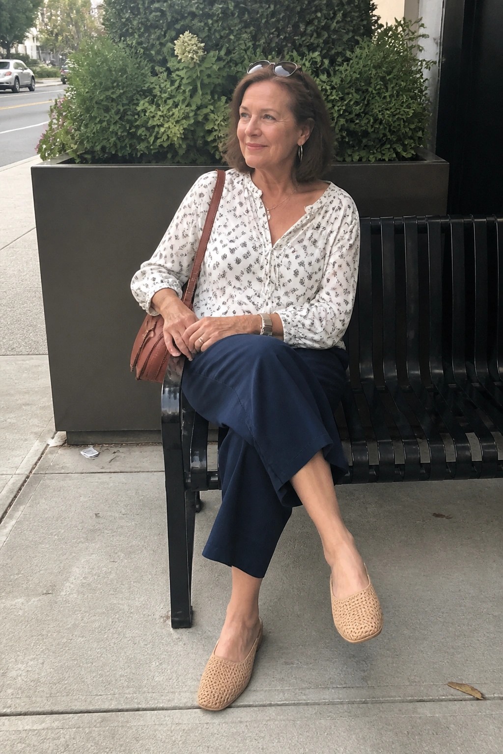 Woman in white patterned blouse and blue pants seated on outdoor black bench with bag