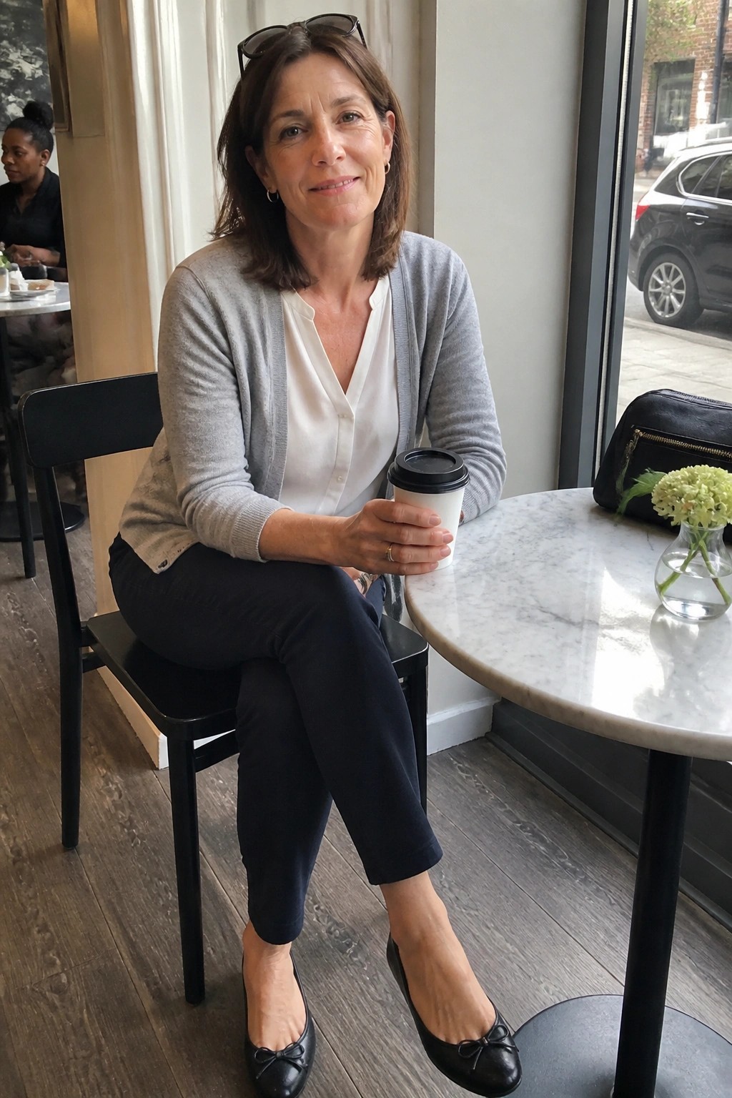 Woman in gray cardigan holds coffee at marble cafe table with flowers nearby
