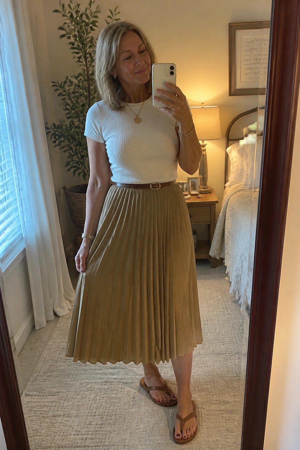 Woman in white top and tan pleated skirt taking mirror selfie in bedroom