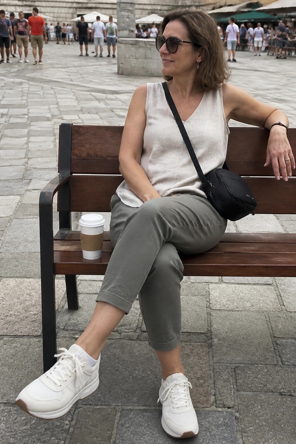 Woman in beige top and olive pants sits on wooden bench with coffee cup