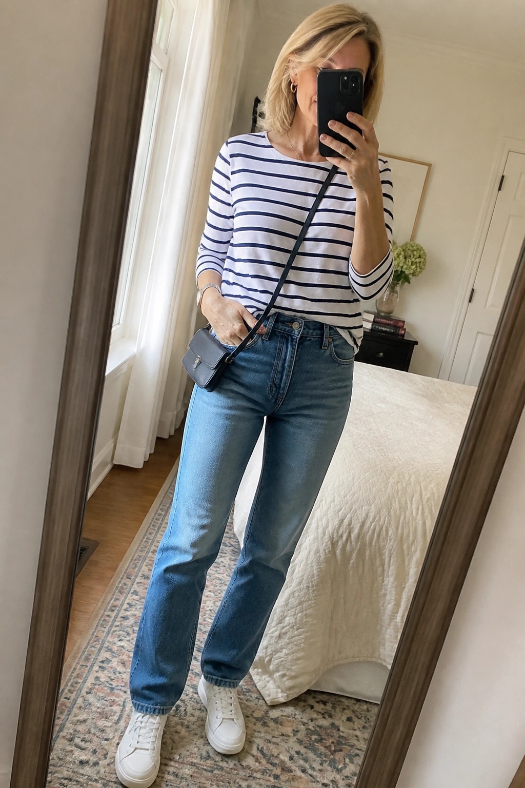 Blonde woman in striped shirt and jeans taking mirror selfie in bedroom