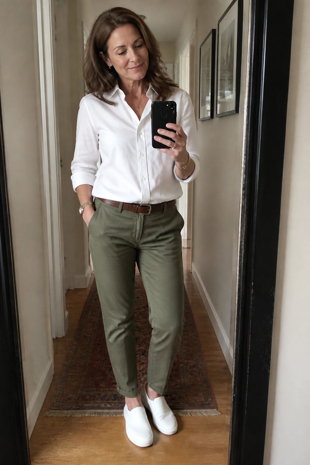 Woman in white shirt and olive pants taking mirror selfie in hallway