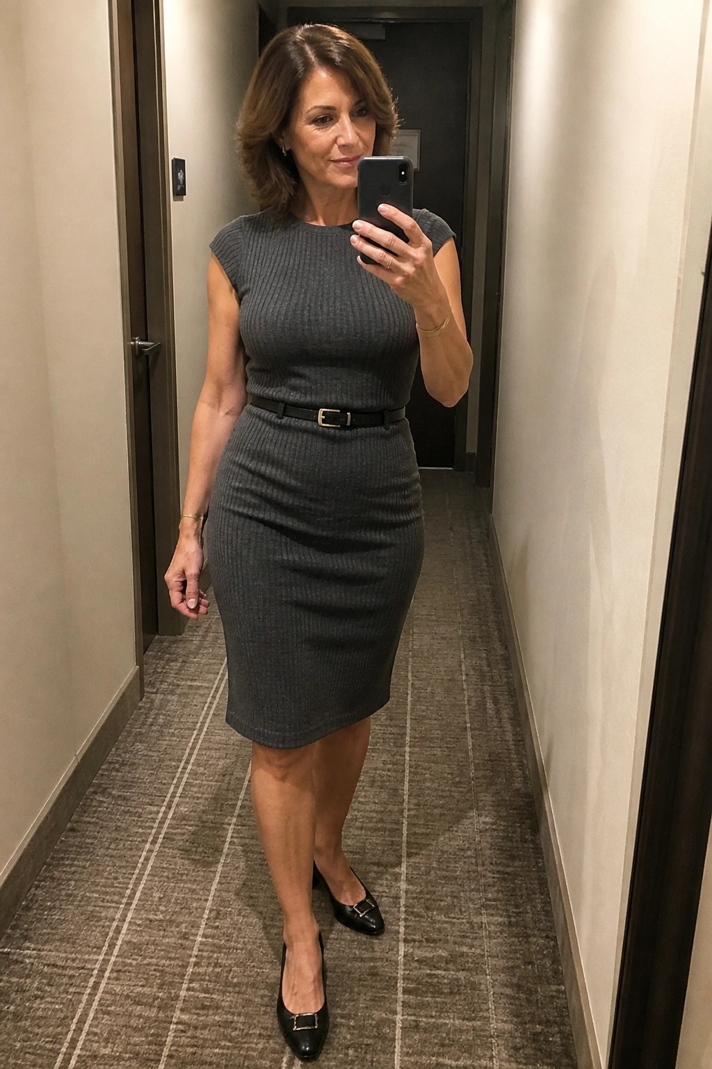 Woman in gray ribbed dress with black belt taking mirror selfie in hallway