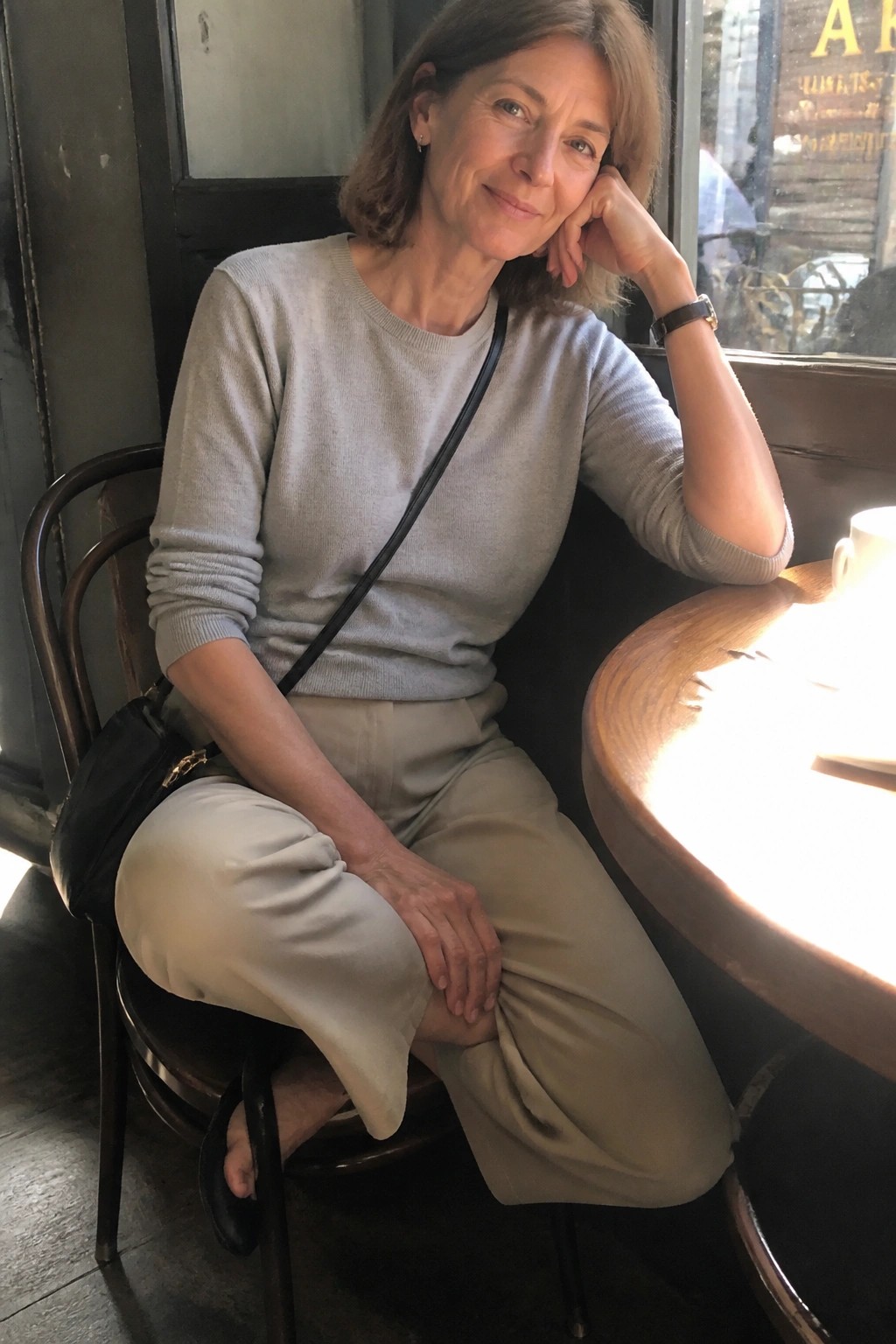 Woman over 50 wearing a light gray knit sweater tucked into beige wide leg pants with a black shoulder bag featuring gold hardware and a black watch on her wrist.