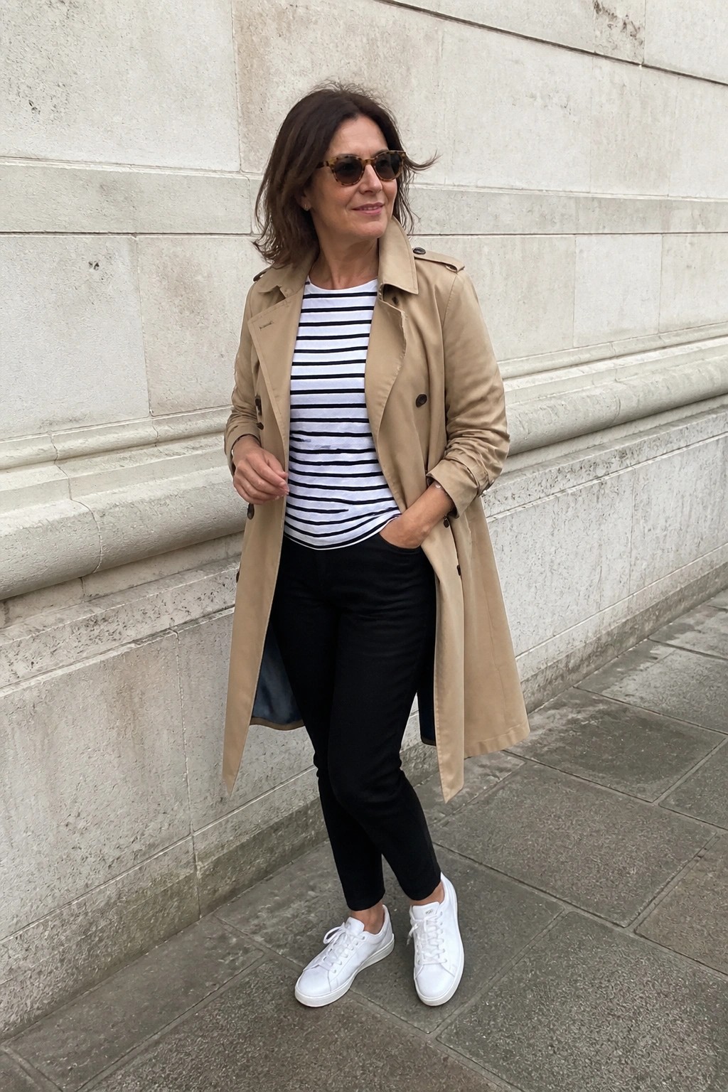 Woman in beige trench coat, striped top, black pants and white sneakers by stone wall.