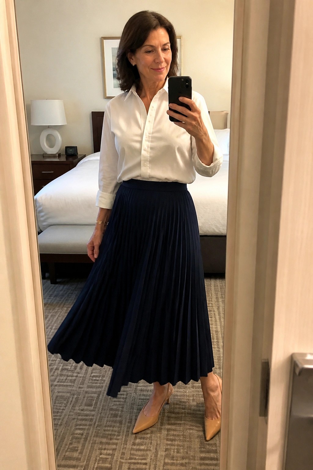 Woman in white shirt and navy pleated skirt taking mirror selfie in bedroom