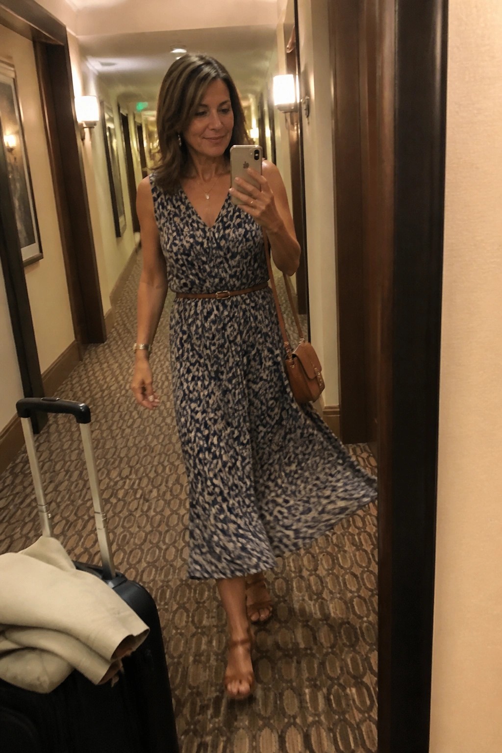 Woman in blue patterned dress taking mirror selfie beside suitcase in hotel hallway