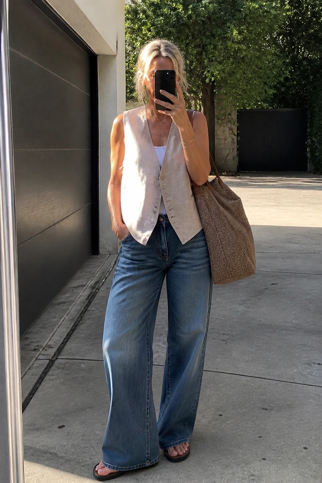 Blonde woman in beige vest and wide-leg jeans taking selfie with tote bag outdoors