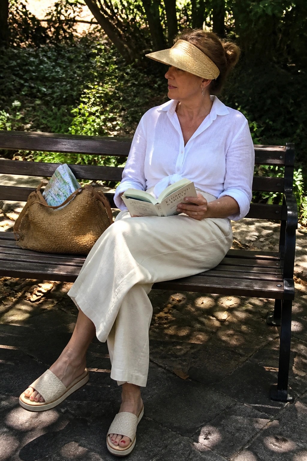 Woman in white shirt and beige pants reading book on park bench wearing straw visor.