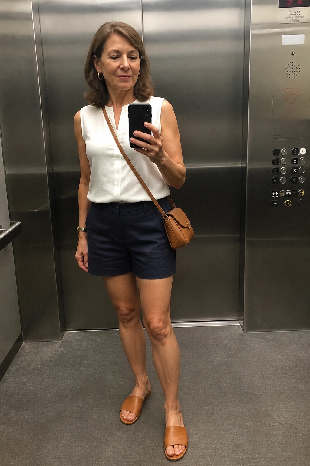 Woman in white top and navy shorts taking selfie in elevator with brown bag