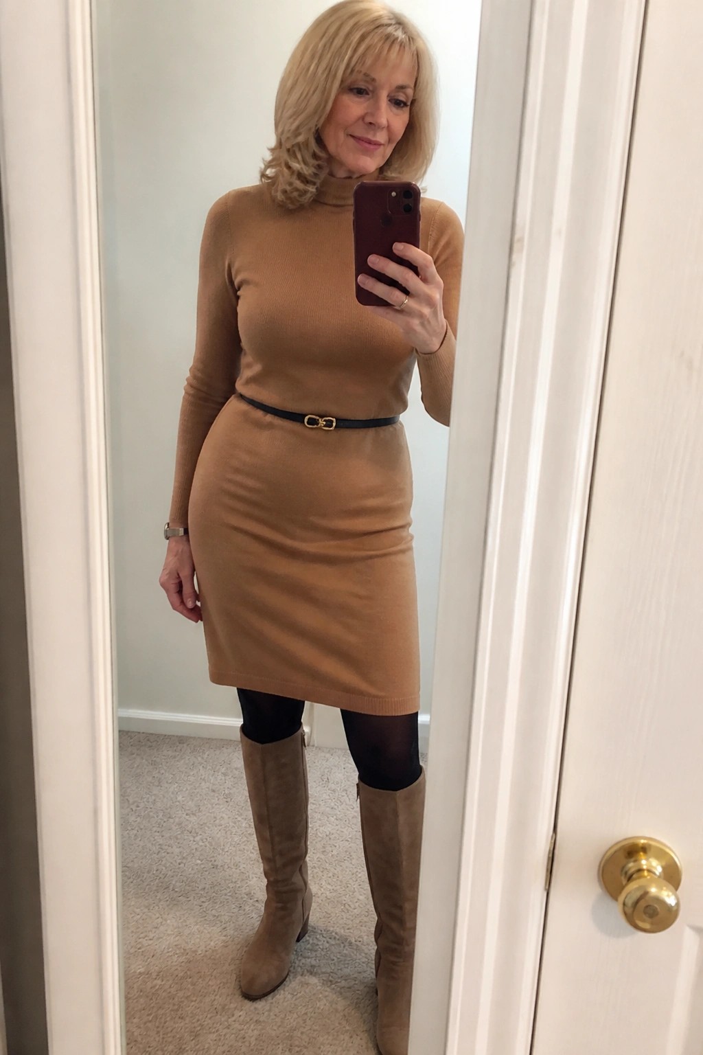 Blonde woman in tan turtleneck sweater dress, black belt, and suede boots taking mirror selfie