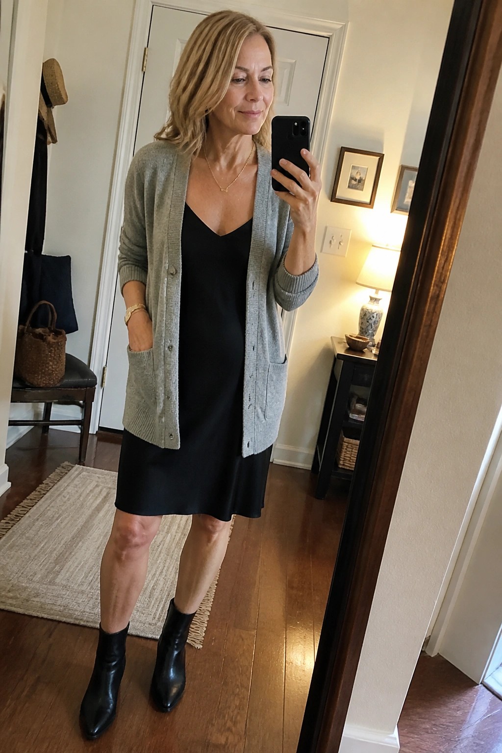 Woman in gray cardigan over black dress taking mirror selfie with phone