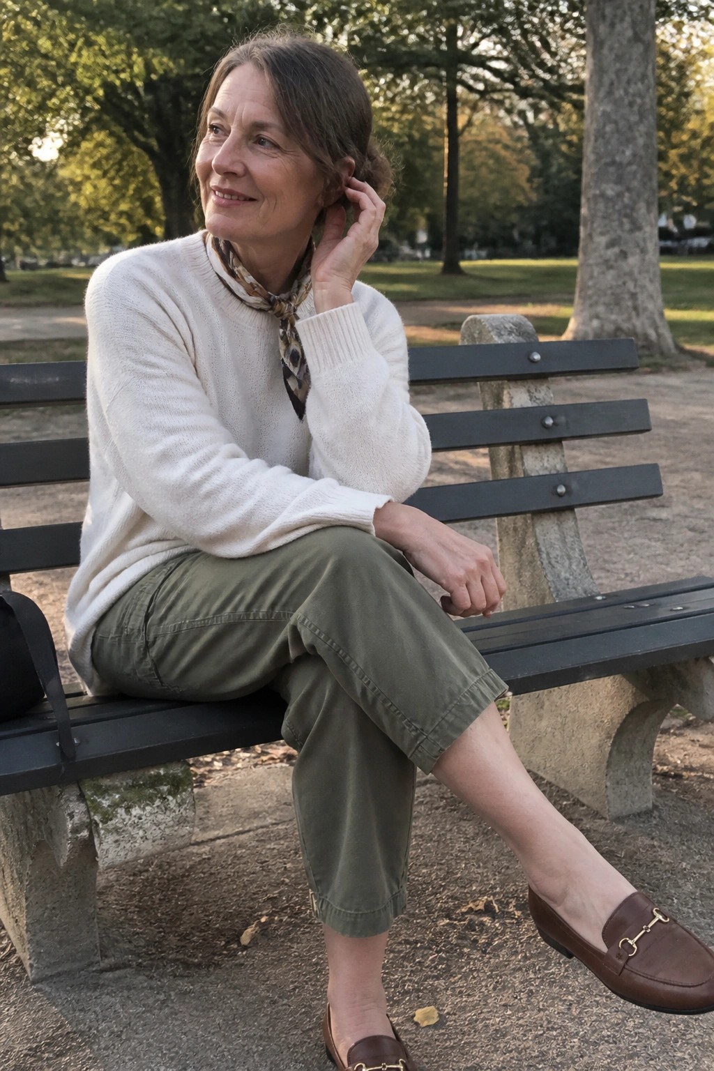 Woman in cream sweater and green pants on park bench, hand to ear.