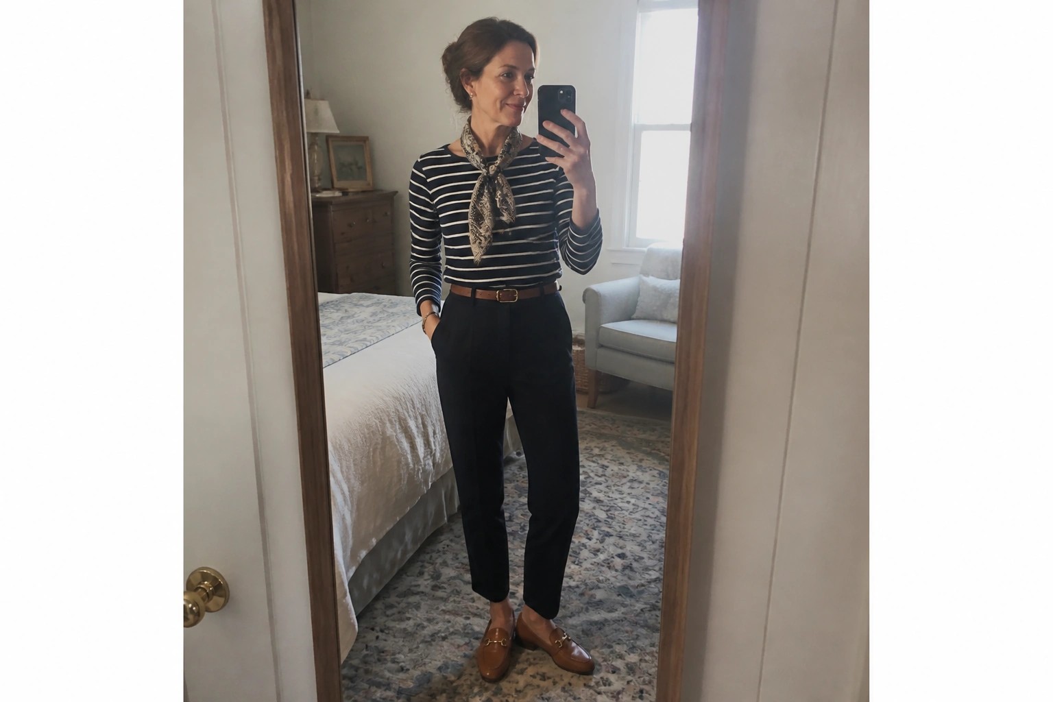 Woman in striped shirt, scarf, black pants, and brown loafers taking mirror selfie