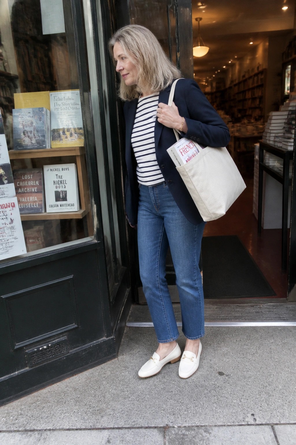 Gray-haired woman in navy blazer and striped shirt with tote bag at bookstore entrance.