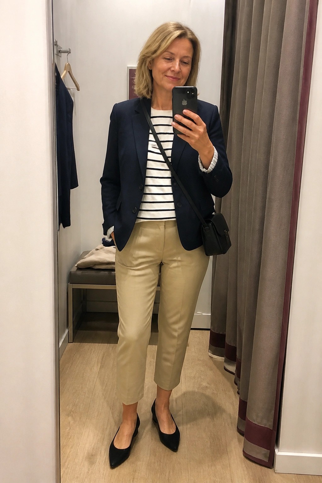 Woman wearing navy blazer over striped top with beige trousers and black pointed flats holding phone for mirror selfie