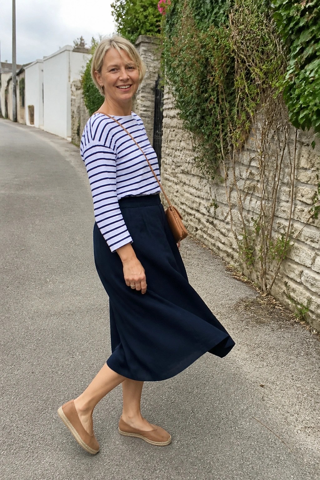 Woman over 50 wearing a blue and white striped long sleeve top tucked into a navy midi skirt with brown espadrille flats and a brown crossbody bag.