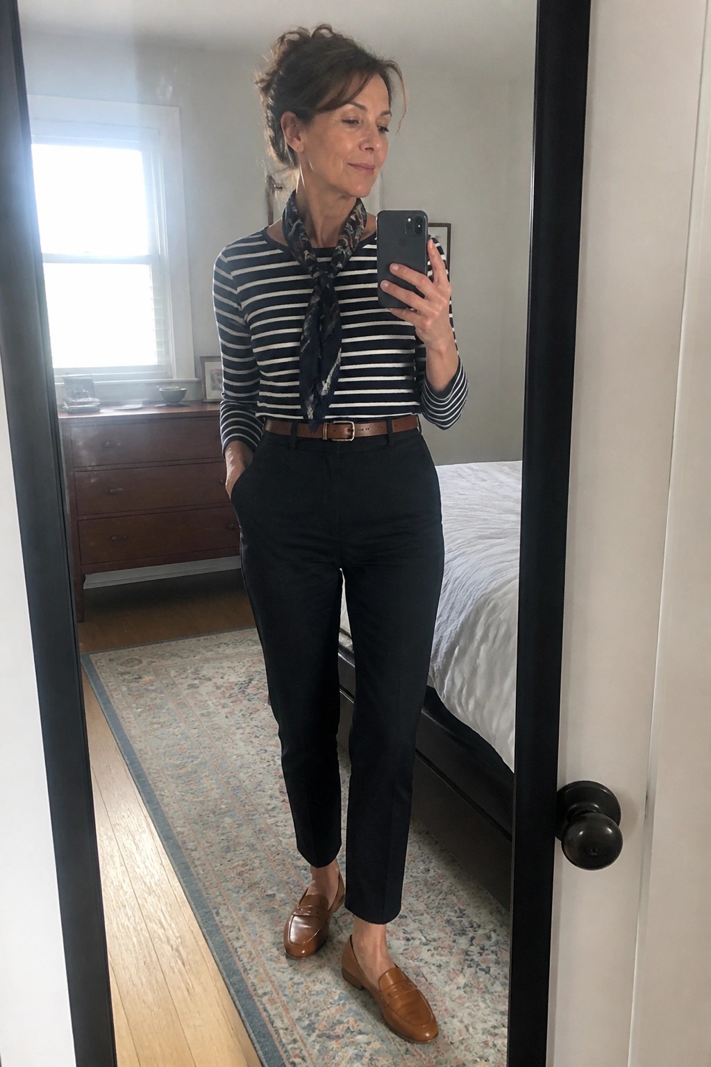 Woman in striped shirt, scarf, black pants, and brown loafers taking mirror selfie