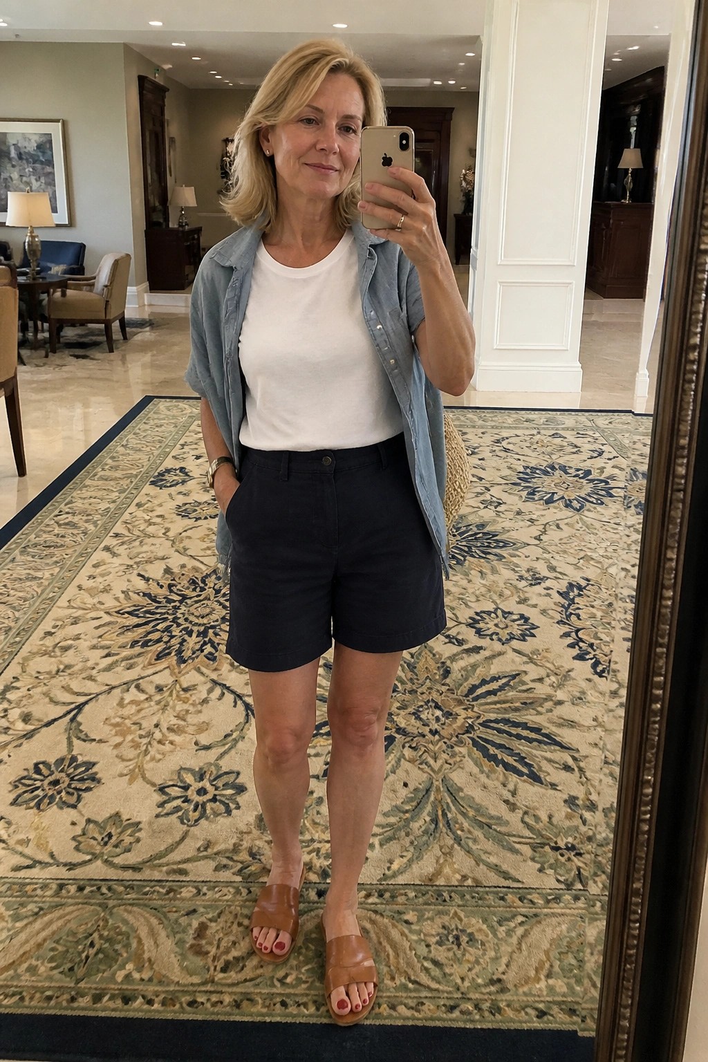 Woman over 50 in mirror selfie wearing white tee under open light blue denim shirt, navy shorts, and brown slide sandals in a hotel lobby setting.