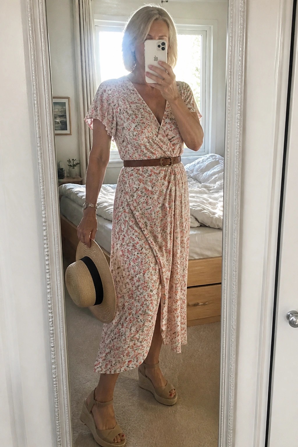Woman in pink floral wrap dress holding phone and straw hat in mirror