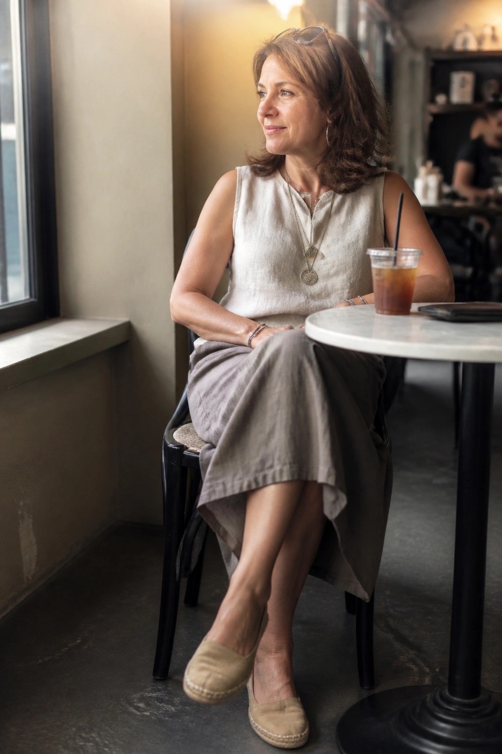 Woman in beige top and gray skirt at cafe table with iced drink
