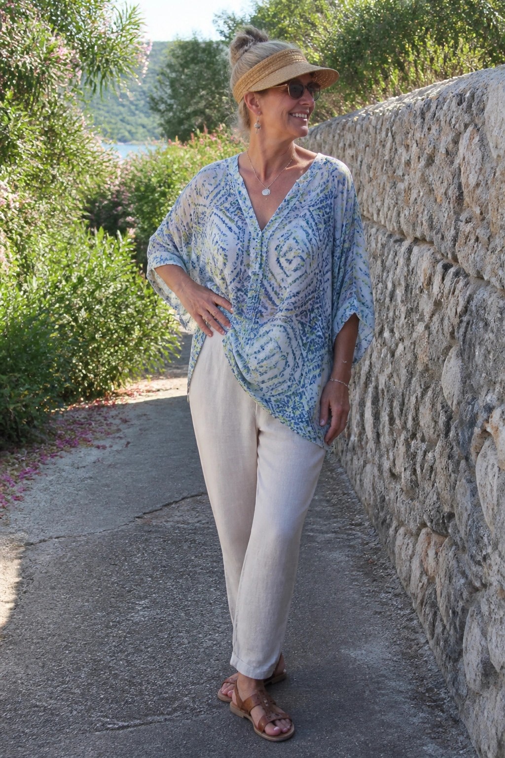 Woman over 50 wearing a loose blue green patterned top over straight beige pants with a straw visor brown sandals and layered necklaces standing beside a stone wall.